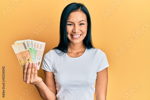 Beautiful hispanic woman holding south korean won banknotes looking positive and happy standing and smiling with a confident smile showing teeth