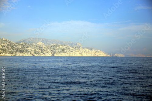 Rocky coast, in the fog, seen from the sea with a small white sailboat, in the distance, between blue sky and sea, Parc National des Calanques, Marseille, France