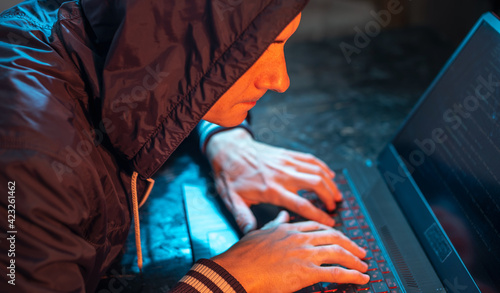 Hooded hacker is typing on a laptop keyboard in a dark room under a neon light. Cybercrime fraud and identity theft