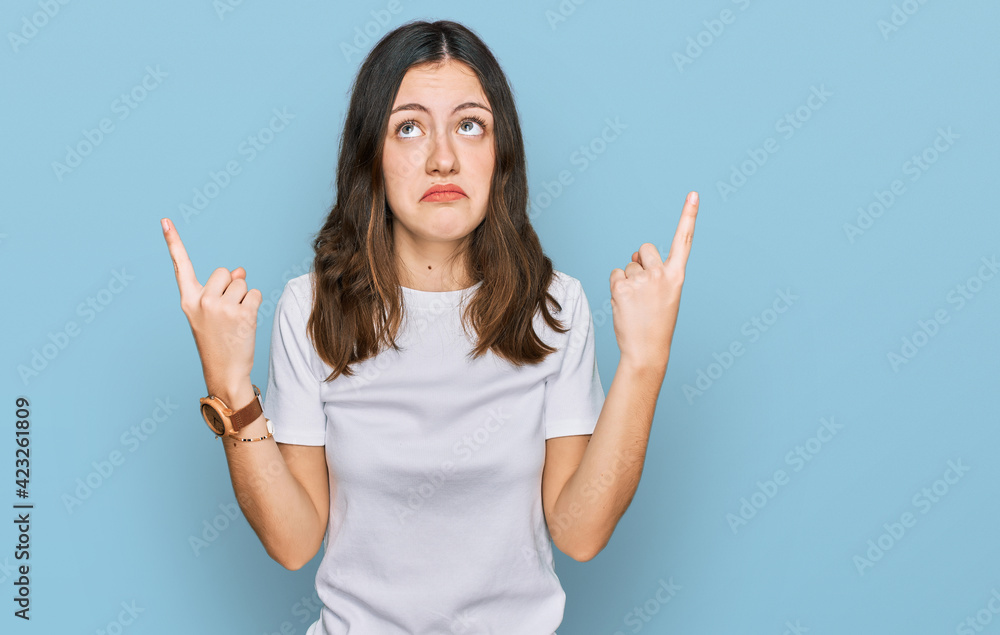 Young beautiful woman wearing casual white t shirt pointing up looking sad and upset, indicating direction with fingers, unhappy and depressed. Young beautiful woman wearing casual white t shirt pointing up looking sad and upset, indicating direction with fingers, unhappy and depressed.