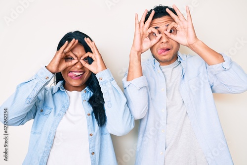 Beautiful latin young couple wearing casual clothes doing ok gesture like binoculars sticking tongue out, eyes looking through fingers. crazy expression.