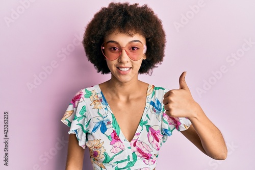 Young hispanic girl wearing summer style and hear sunglasses smiling happy and positive, thumb up doing excellent and approval sign
