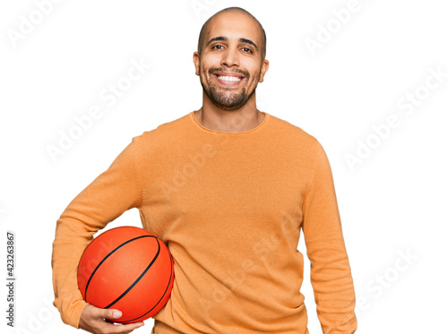Hispanic adult man holding basketball ball looking positive and happy standing and smiling with a confident smile showing teeth