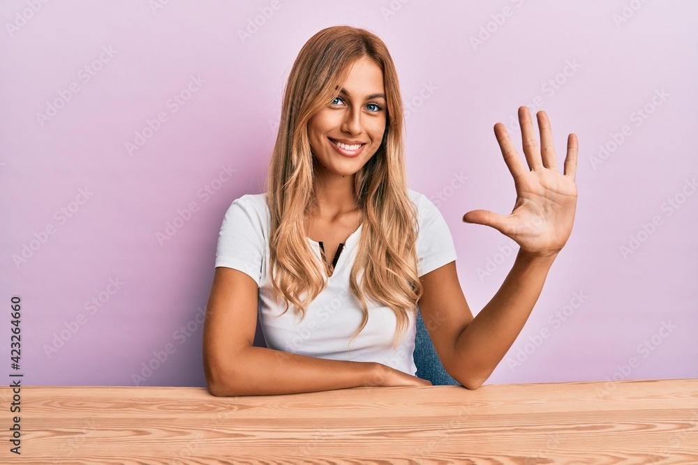 Beautiful blonde young woman wearing casual clothes sitting on the table showing and pointing up with fingers number five while smiling confident and happy. Beautiful blonde young woman wearing casual clothes sitting on the table showing and pointing up with fingers number five while smiling confident and happy.
