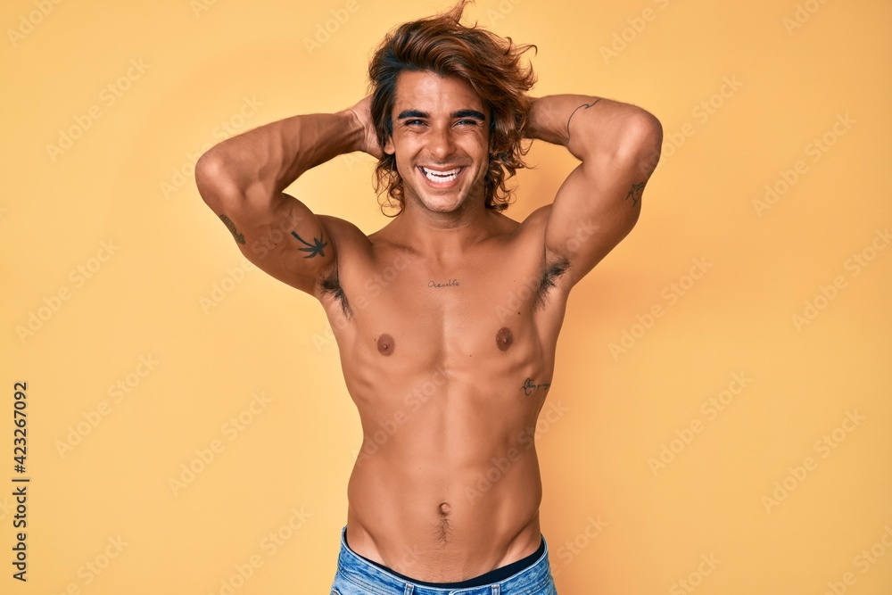 Young hispanic man standing shirtless relaxing and stretching, arms and hands behind head and neck smiling happy