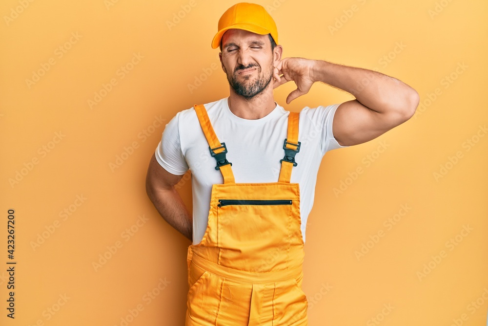 Young handsome man wearing handyman uniform over yellow background suffering of neck ache injury, touching neck with hand, muscular pain