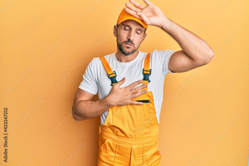 Young handsome man wearing handyman uniform over yellow background touching forehead for illness and fever, flu and cold, virus sick