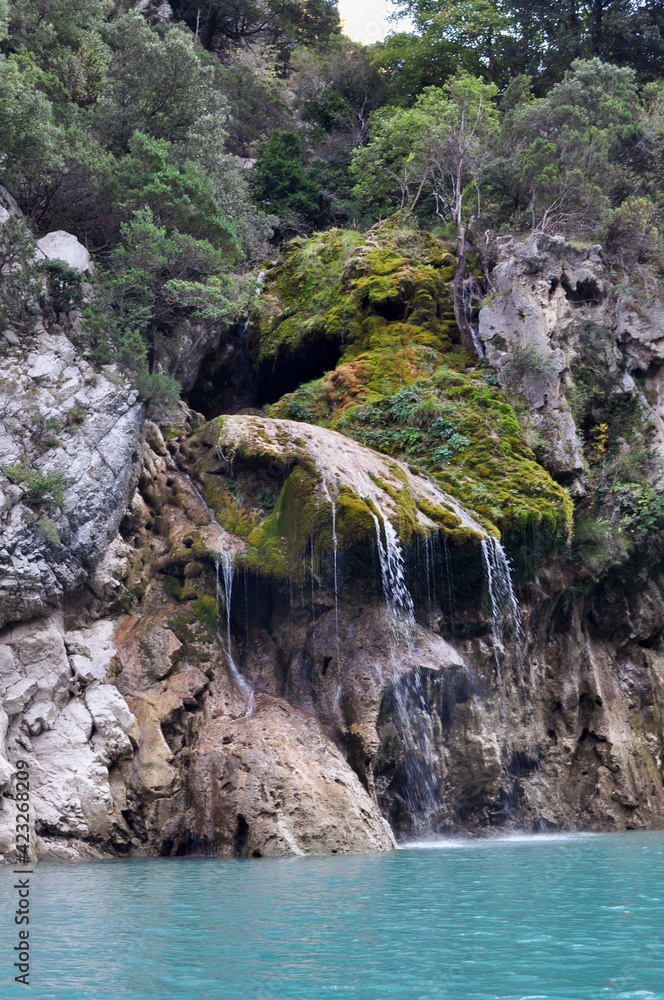 A rock in the French Mountains with a small waterfall flowing into the clear turquoise water - Lake Lac de Sainte-Croix, France A rock in the French Mountains with a small waterfall flowing into the clear turquoise water - Lake Lac de Sainte-Croix, France