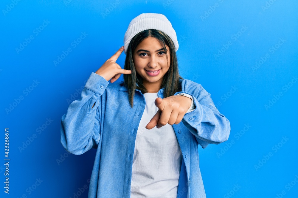 Young latin woman wearing cute wool cap smiling doing talking on the telephone gesture and pointing to you. call me.