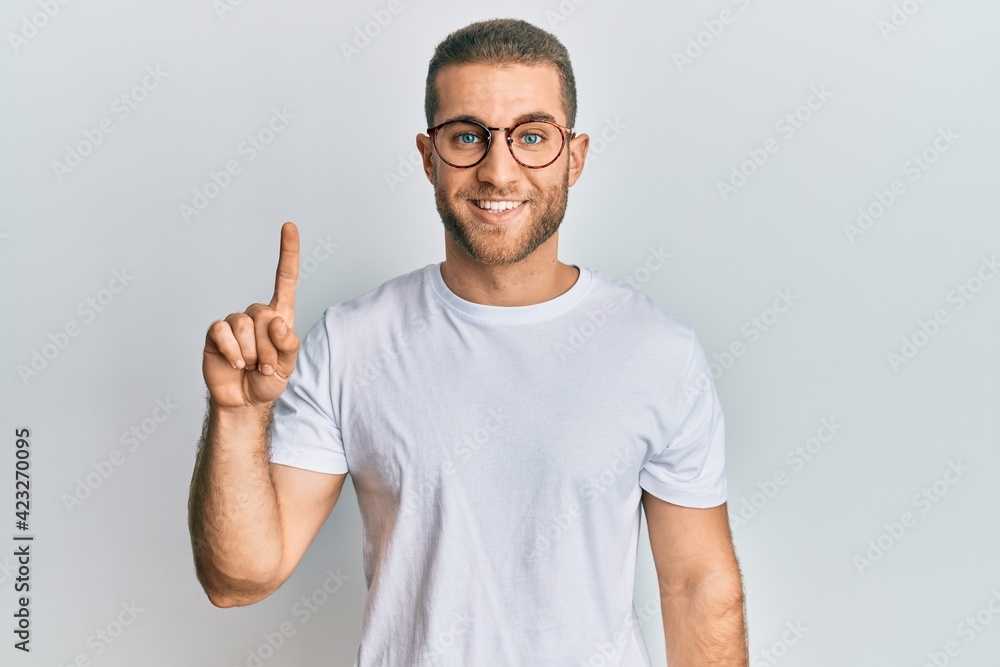 Young caucasian man wearing casual clothes and glasses showing and pointing up with finger number one while smiling confident and happy. Young caucasian man wearing casual clothes and glasses showing and pointing up with finger number one while smiling confident and happy.