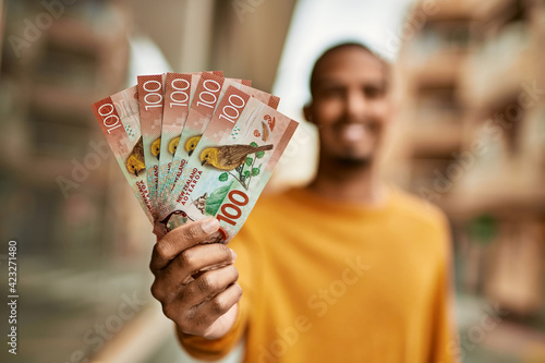 Young african american man smiling happy holding new zealand dollars at the city.