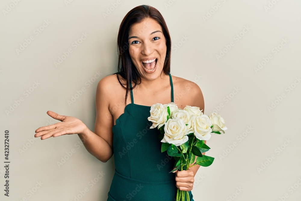 Young latin woman holding flowers celebrating achievement with happy smile and winner expression with raised hand Young latin woman holding flowers celebrating achievement with happy smile and winner expression with raised hand