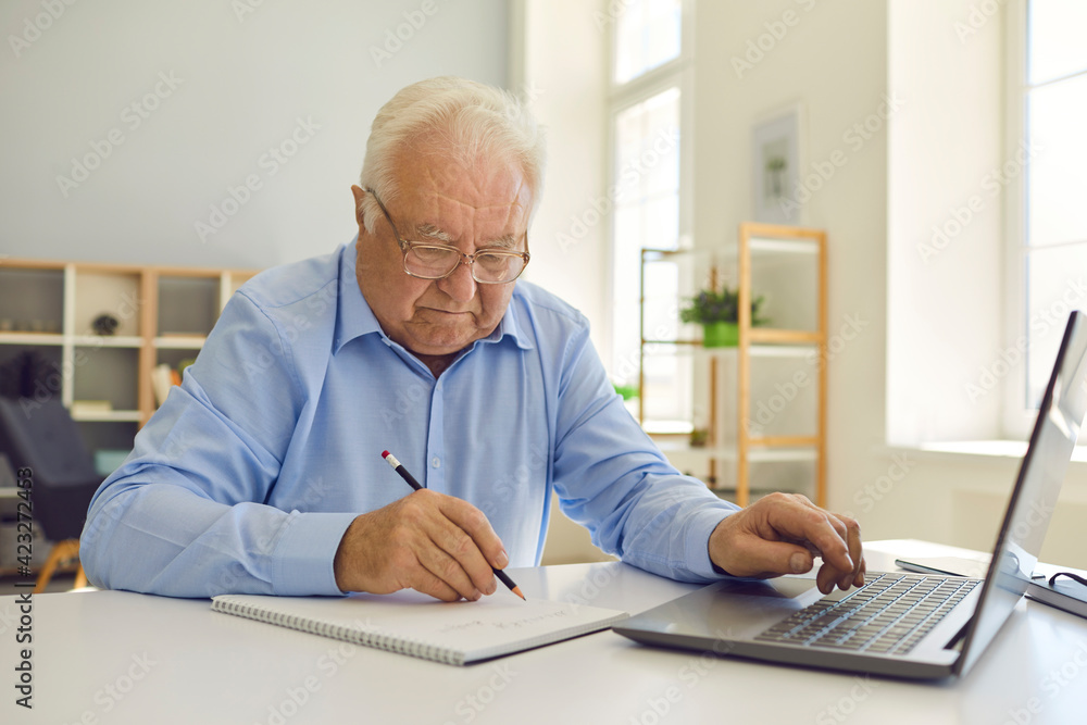 Senior man in eyeglasses sitting at desk at home, using laptop and taking notes in notebook Senior man in eyeglasses sitting at desk at home, using laptop and taking notes in notebook