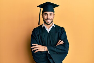 Young hispanic man wearing graduation cap and ceremony robe happy face smiling with crossed arms looking at the camera. positive person.