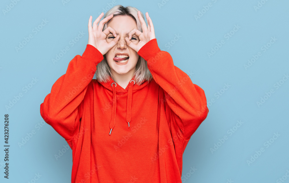Young blonde girl wearing casual clothes doing ok gesture like binoculars sticking tongue out, eyes looking through fingers. crazy expression. Young blonde girl wearing casual clothes doing ok gesture like binoculars sticking tongue out, eyes looking through fingers. crazy expression.
