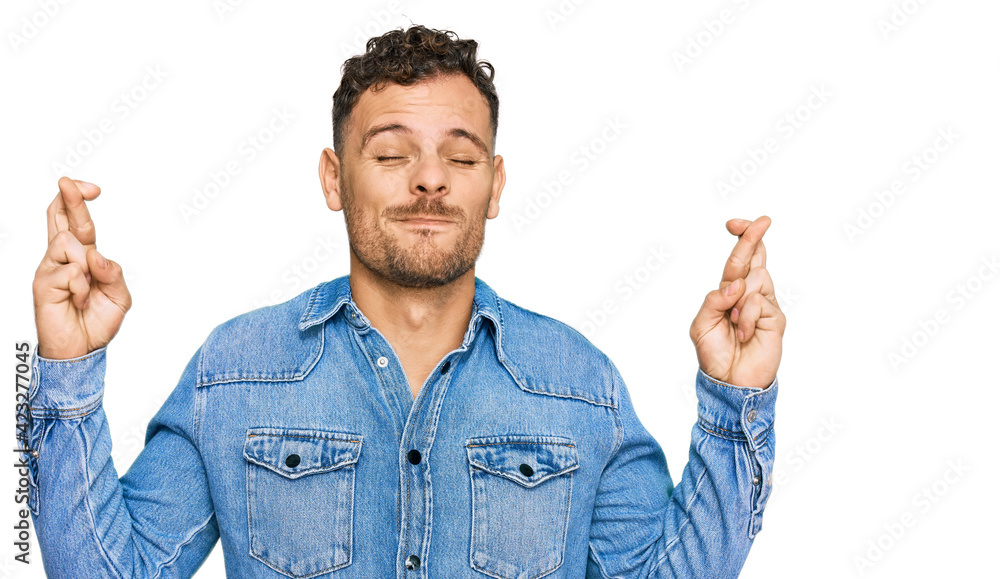 Young hispanic man wearing casual denim jacket gesturing finger crossed smiling with hope and eyes closed. luck and superstitious concept. Young hispanic man wearing casual denim jacket gesturing finger crossed smiling with hope and eyes closed. luck and superstitious concept.