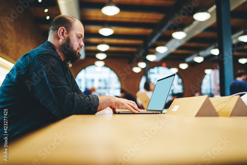 Side view of skilled male freelancer with beard using mock up laptop computer with copy space area for internet advertising, software developer updating data programs during online remote work