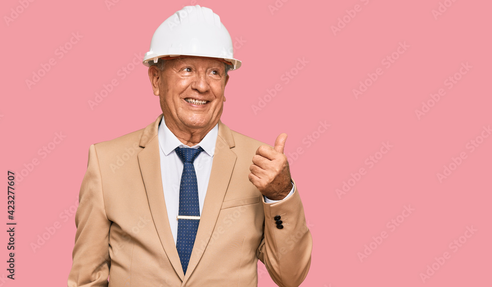 Senior caucasian man wearing architect hardhat smiling with happy face looking and pointing to the side with thumb up. Senior caucasian man wearing architect hardhat smiling with happy face looking and pointing to the side with thumb up.