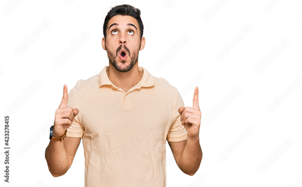Young hispanic man wearing casual clothes amazed and surprised looking up and pointing with fingers and raised arms. Young hispanic man wearing casual clothes amazed and surprised looking up and pointing with fingers and raised arms.