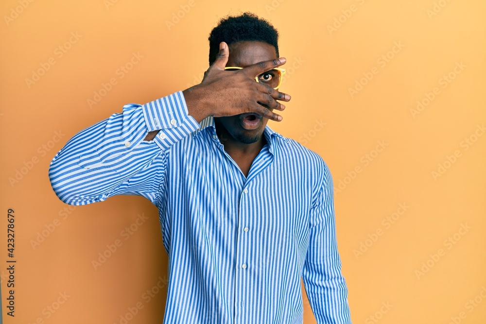 Handsome black man wearing yellow glasses peeking in shock covering face and eyes with hand, looking through fingers afraid Handsome black man wearing yellow glasses peeking in shock covering face and eyes with hand, looking through fingers afraid