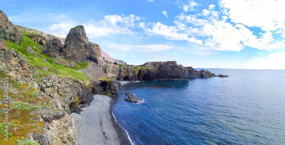 One person standing in scenic view in a distance on the rock near Blue ocean with black sand beach and green coastline with rocks and cliffs and blue cloudy sky