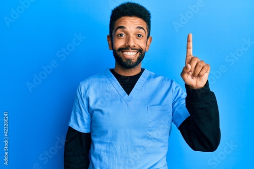 Handsome hispanic man with beard wearing blue male nurse uniform pointing finger up with successful idea. exited and happy. number one.