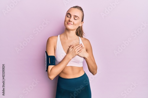 Beautiful blonde woman wearing sportswear and arm band smiling with hands on chest with closed eyes and grateful gesture on face. health concept.