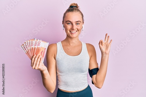 Beautiful blonde sport woman holding 100 norwegian krone banknotes doing ok sign with fingers, smiling friendly gesturing excellent symbol
