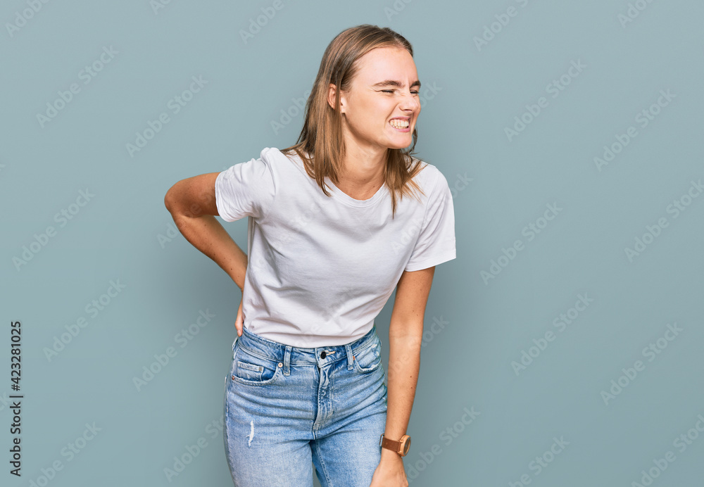 Beautiful young blonde woman wearing casual white t shirt suffering of backache, touching back with hand, muscular pain Beautiful young blonde woman wearing casual white t shirt suffering of backache, touching back with hand, muscular pain