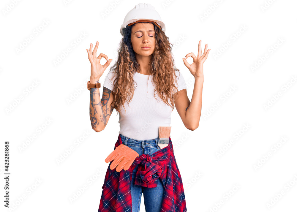 Young hispanic woman with tattoo wearing hardhat and builder clothes relax and smiling with eyes closed doing meditation gesture with fingers. yoga concept. Young hispanic woman with tattoo wearing hardhat and builder clothes relax and smiling with eyes closed doing meditation gesture with fingers. yoga concept.