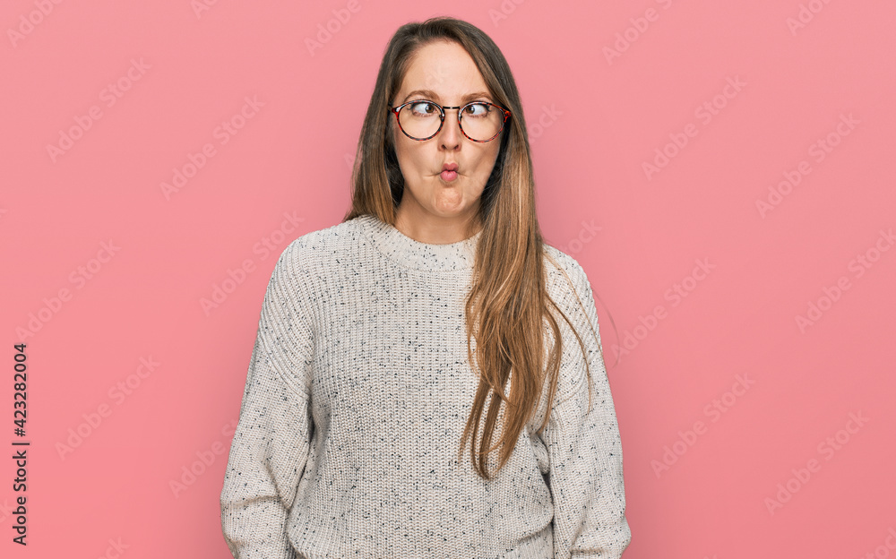 Young blonde woman wearing casual sweater and glasses making fish face with lips, crazy and comical gesture. funny expression.