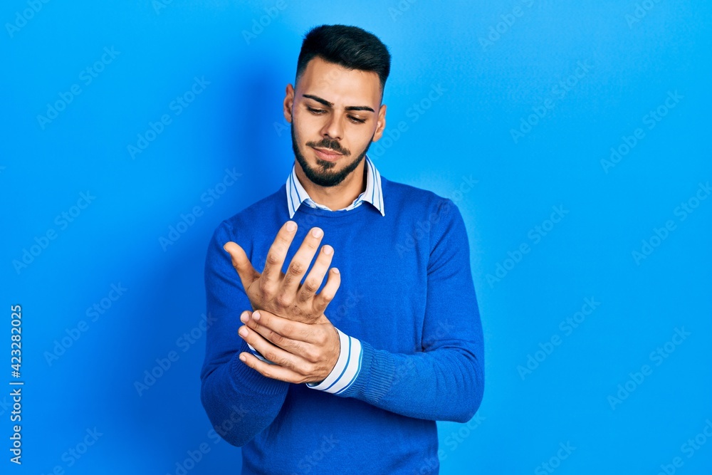 Young hispanic man with beard wearing casual blue sweater suffering pain on hands and fingers, arthritis inflammation Young hispanic man with beard wearing casual blue sweater suffering pain on hands and fingers, arthritis inflammation