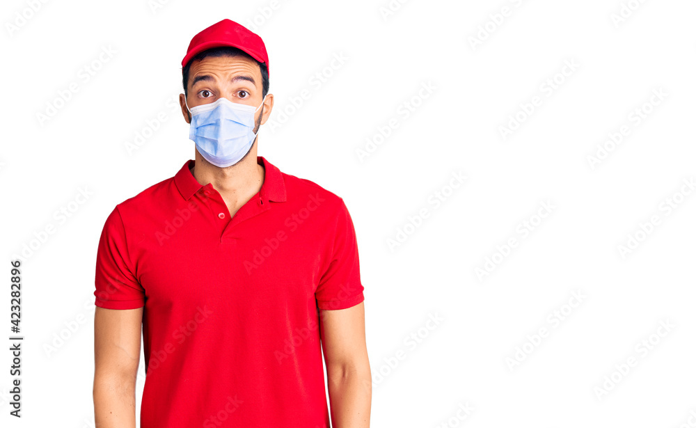 Young handsome hispanic man wearing delivery uniform and medical mask scared and amazed with open mouth for surprise, disbelief face Young handsome hispanic man wearing delivery uniform and medical mask scared and amazed with open mouth for surprise, disbelief face