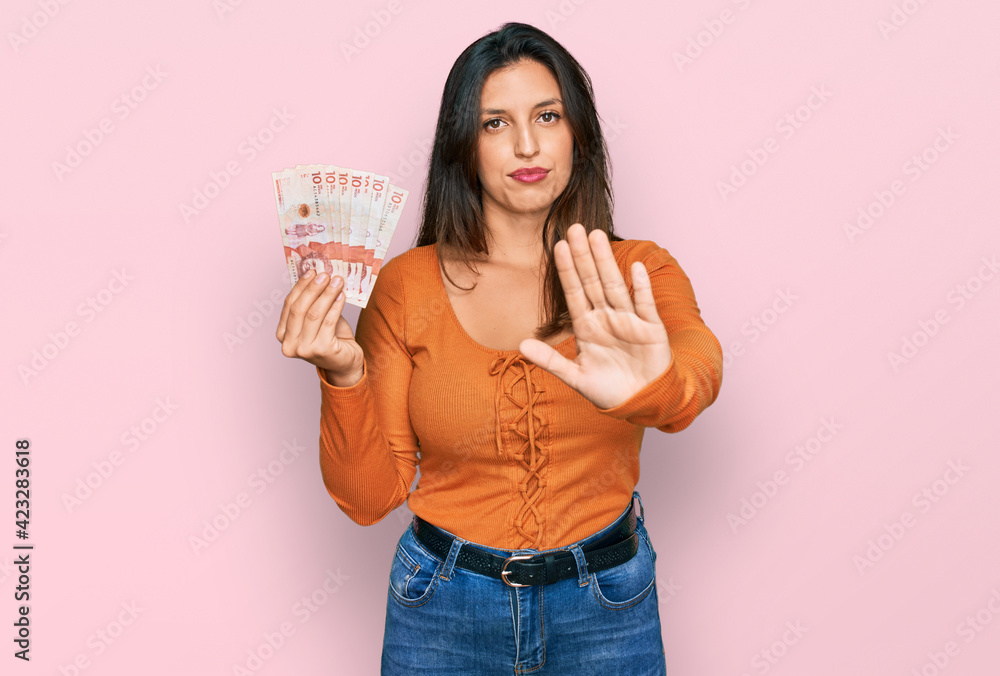 Beautiful hispanic woman holding 10 colombian pesos banknotes with open hand doing stop sign with serious and confident expression, defense gesture Beautiful hispanic woman holding 10 colombian pesos banknotes with open hand doing stop sign with serious and confident expression, defense gesture
