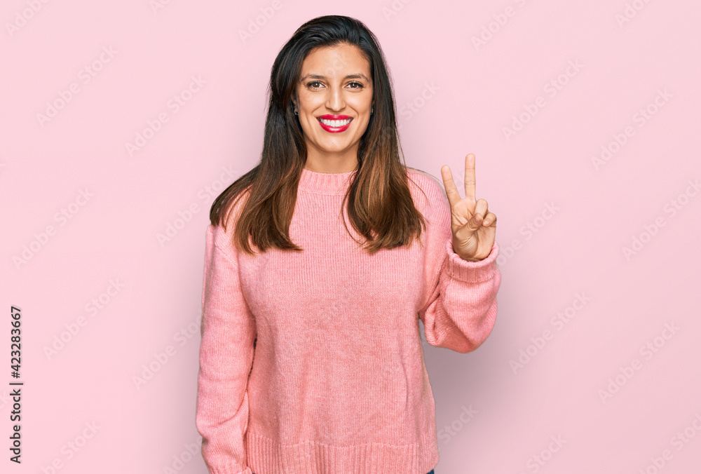 Beautiful hispanic woman wearing casual winter sweater smiling with happy face winking at the camera doing victory sign. number two. Beautiful hispanic woman wearing casual winter sweater smiling with happy face winking at the camera doing victory sign. number two.
