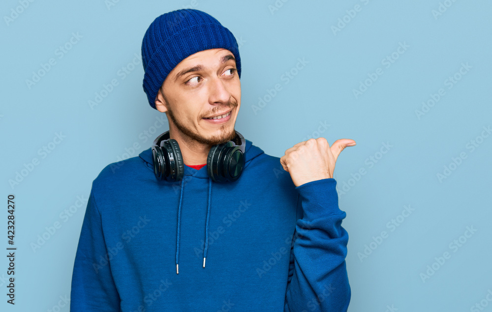 Hispanic young man wearing sweatshirt and wool hat smiling with happy face looking and pointing to the side with thumb up. Hispanic young man wearing sweatshirt and wool hat smiling with happy face looking and pointing to the side with thumb up.