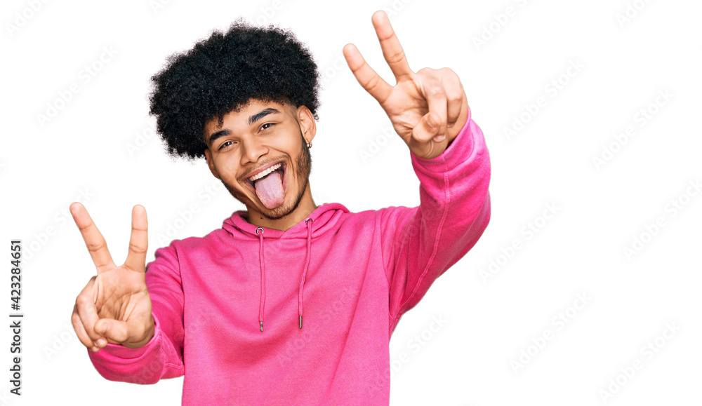 Young african american man with afro hair wearing casual pink sweatshirt smiling with tongue out showing fingers of both hands doing victory sign. number two. Young african american man with afro hair wearing casual pink sweatshirt smiling with tongue out showing fingers of both hands doing victory sign. number two.