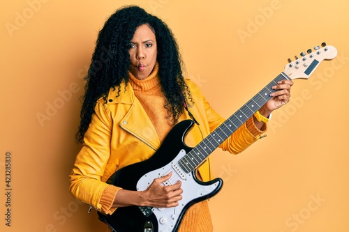 Middle age african american woman playing electric guitar making fish face with mouth and squinting eyes, crazy and comical.