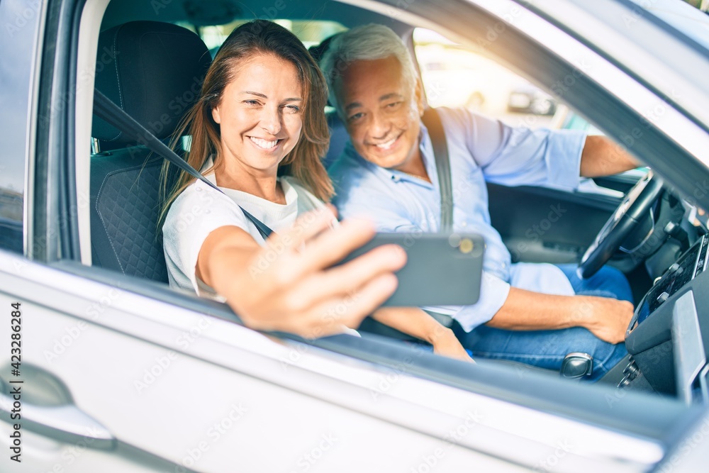 Middle age couple in love sitting inside the car going for a trip taking a selfie picture with smartphone smiling happy and cheerful together