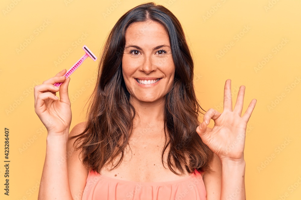 Obraz premium Beautiful brunette woman asking for depilation holding shaver razor over yellow background doing ok sign with fingers, smiling friendly gesturing excellent symbol
