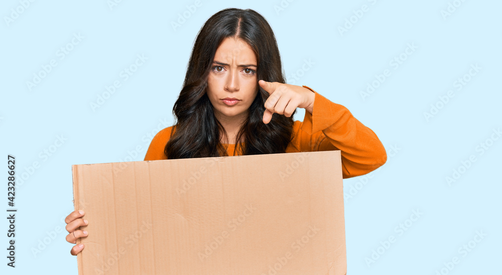 Beautiful brunette young woman holding cardboard blank empty banner pointing with finger to the camera and to you, confident gesture looking serious Beautiful brunette young woman holding cardboard blank empty banner pointing with finger to the camera and to you, confident gesture looking serious