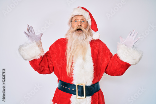 Old senior man with grey hair and long beard wearing santa claus costume with suspenders celebrating crazy and amazed for success with arms raised and open eyes screaming excited. winner concept
