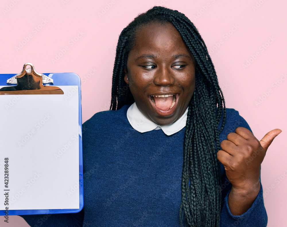 Young black woman with braids holding clipboard with blank space pointing thumb up to the side smiling happy with open mouth Young black woman with braids holding clipboard with blank space pointing thumb up to the side smiling happy with open mouth