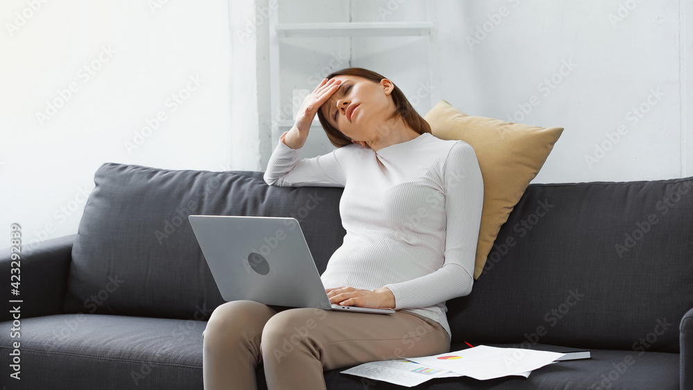 exhausted, pregnant woman suffering from headache near laptop and papers on sofa exhausted, pregnant woman suffering from headache near laptop and papers on sofa
