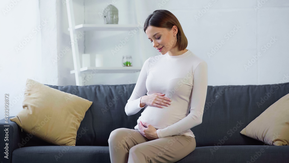 smiling, pregnant woman touching tummy while sitting on sofa at home