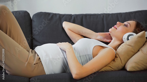 side view of young pregnant woman lying on couch and listening music in headphones