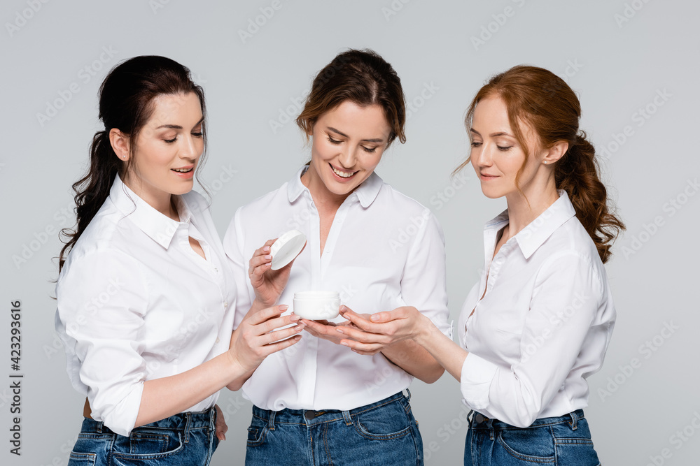 Smiling women looking at jar with cosmetic cream near friend isolated on grey Smiling women looking at jar with cosmetic cream near friend isolated on grey