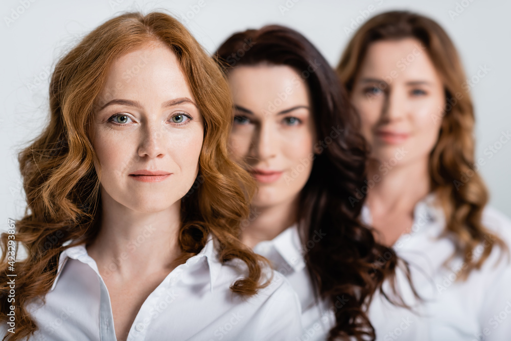 Redhead woman in shirt looking at camera near women on blurred background isolated on grey Redhead woman in shirt looking at camera near women on blurred background isolated on grey