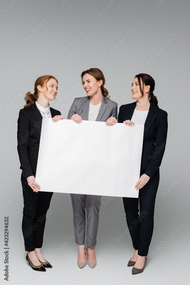 Businesswomen with empty white board smiling at each other on grey background Businesswomen with empty white board smiling at each other on grey background