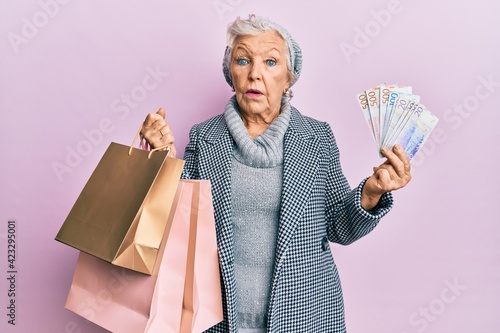 Senior grey-haired woman holding shopping bags and swedish krona banknotes in shock face, looking skeptical and sarcastic, surprised with open mouth